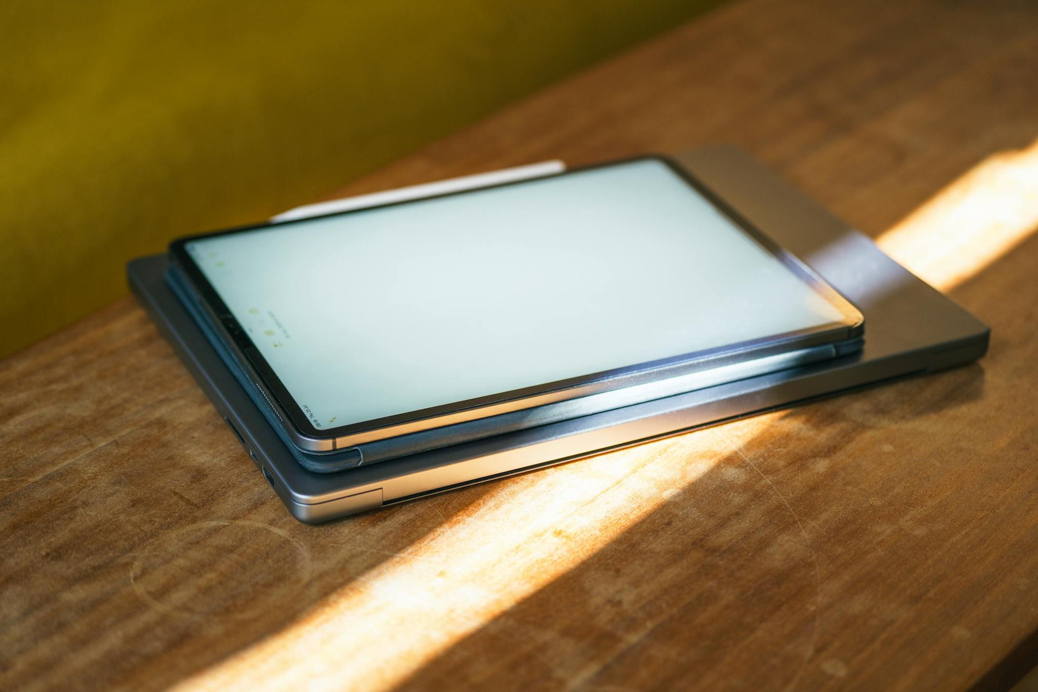 A sleek tablet with stylus atop a laptop on a wooden table, bathed in sunlight.