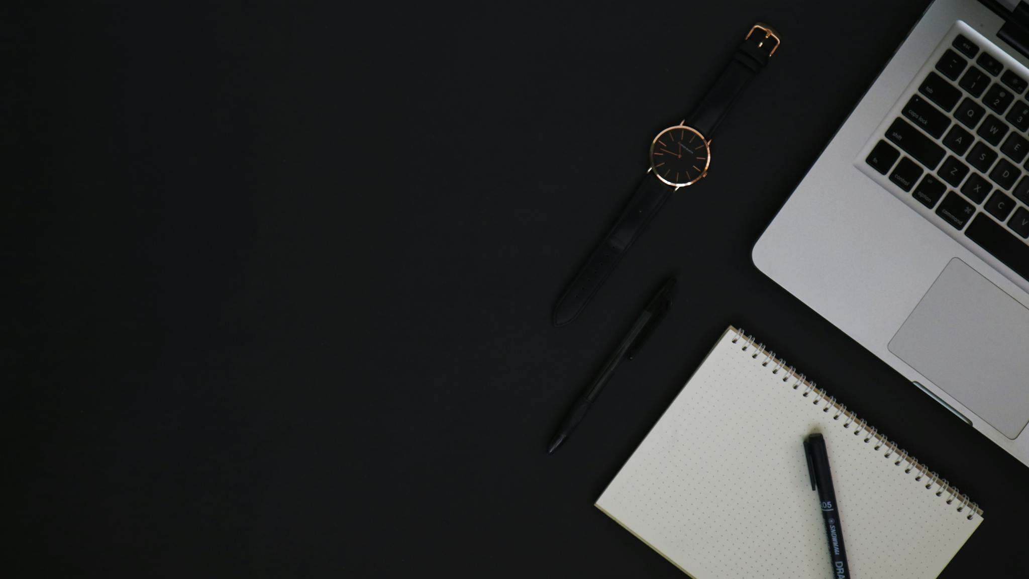 Top-view of a desk setup with a laptop, watch, notebook, and pens on a dark background.