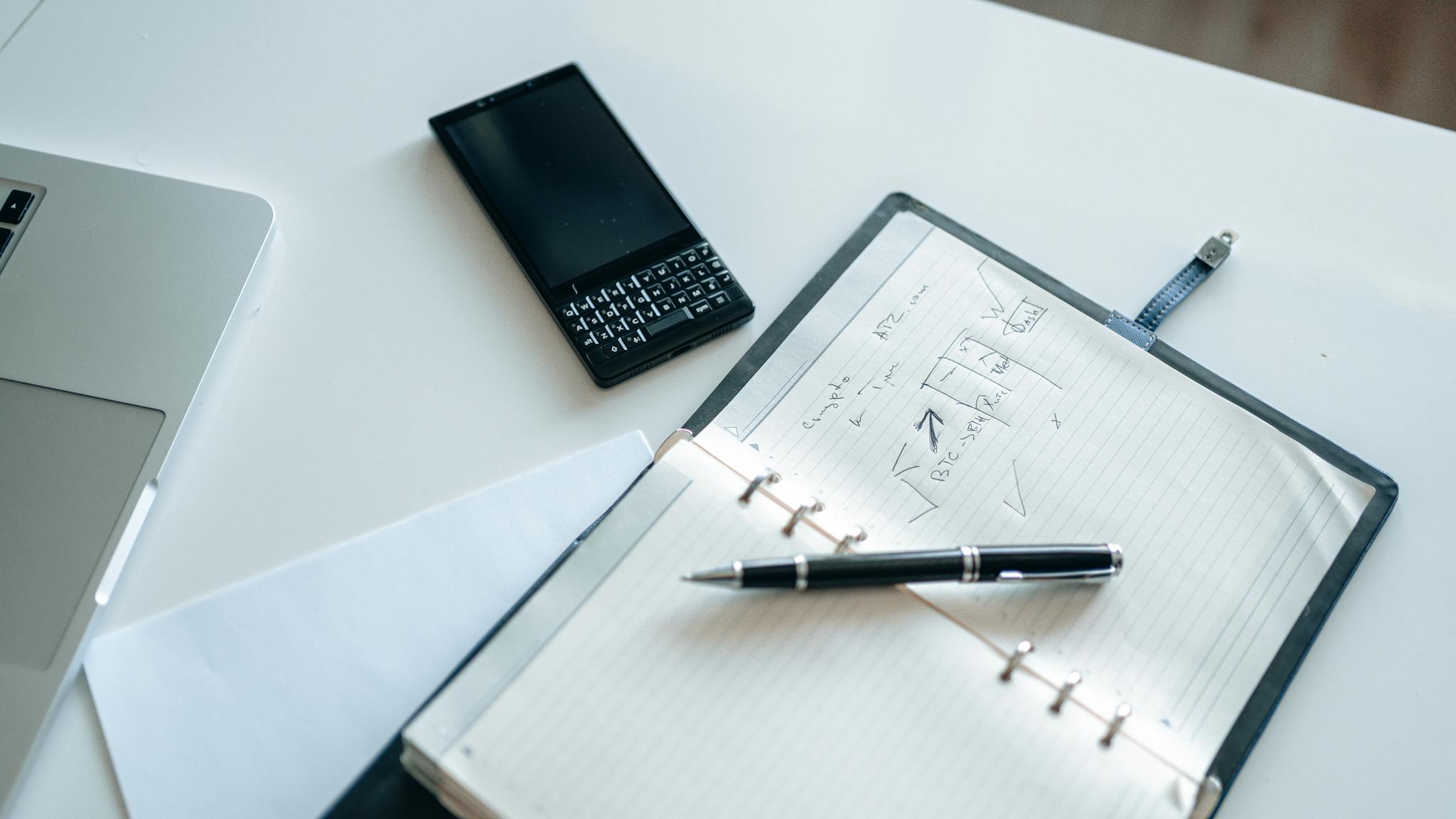 Top view of a notebook, pen, smartphone, and laptop on a desk.